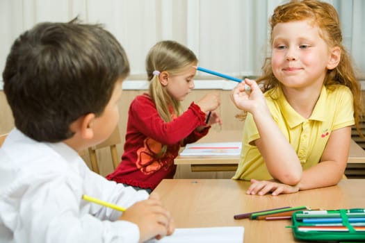 Photo of cute schoolgirl with blue pencil in hand looking at her classmate during lesson