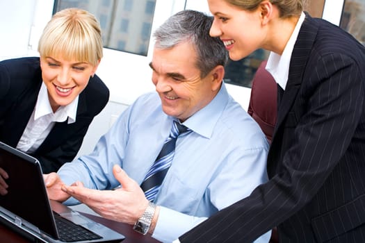 Photo of confident boss pointing at laptop screen and showing something to his young colleagues in the office
