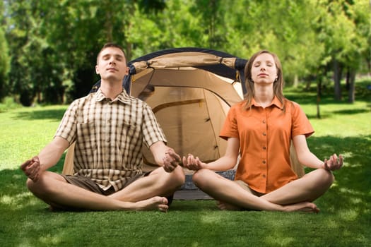 Handsome man and pretty woman meditating on green grass with tent behind