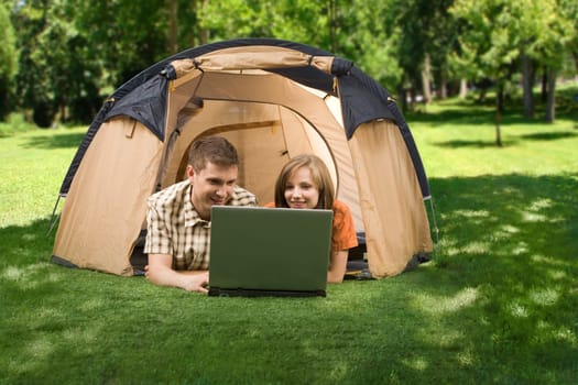 Photo of cheerful girl and guy lying on green grass and looking at laptop monitor in park