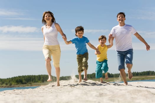 Photo of happy parents holding their sons by hands and running down sandy shore with bright blue sky at background