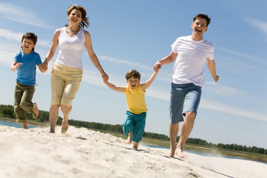 Photo of happy parents holding their sons by hands and running down sandy shore with bright blue sky at background