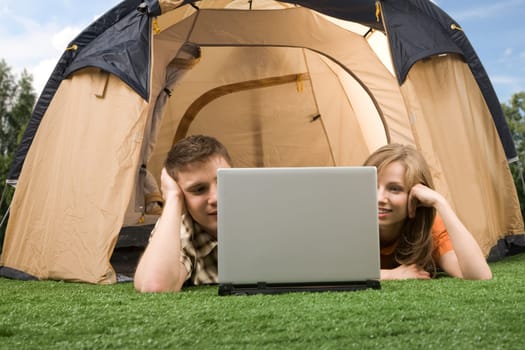 Photo of modern couple lying on green grass and looking at laptop monitor in tent