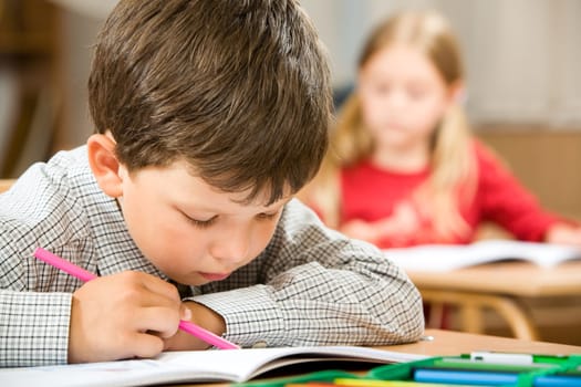 Photo of clever schoolkid writing something in his copybook at lesson on background of classmate