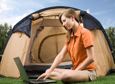 Photo of charming female sitting on green grass and typing with tent near by