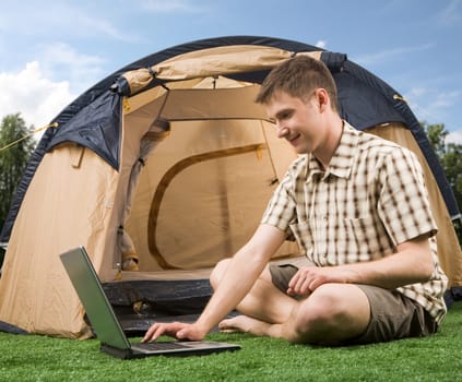 Photo of smiling man sitting on green grass and typing with tent near by