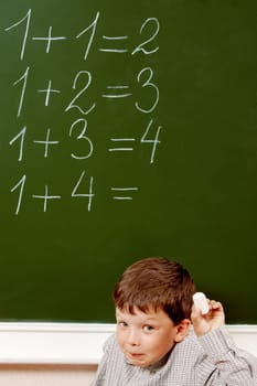 Portrait of curious schoolchild standing at blackboard with written sums on it and looking at camera