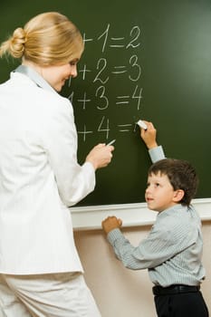 Photo of elementary student doing sums on blackboard and looking at his teacher who is helping him