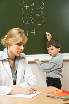 Image of schoolboy writing numbers on the blackboard and teacher sitting at the table  near by