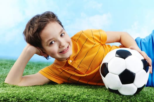 Image of happy boy lying on the grass with ball and looking at camera