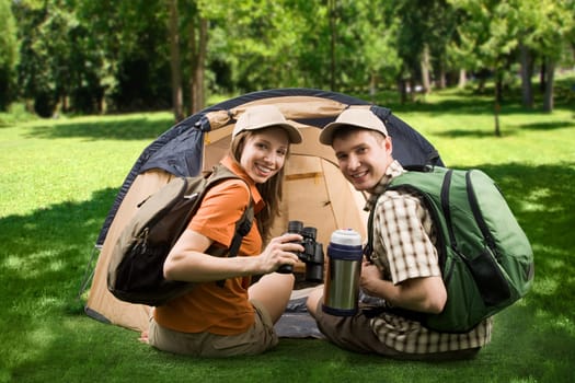 Photo of cheerful girl and guy sitting on green grass by tent and looking at camera with smiles