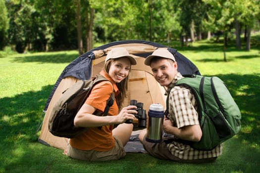Photo of cheerful girl and guy sitting on green grass by tent and looking at camera with smiles