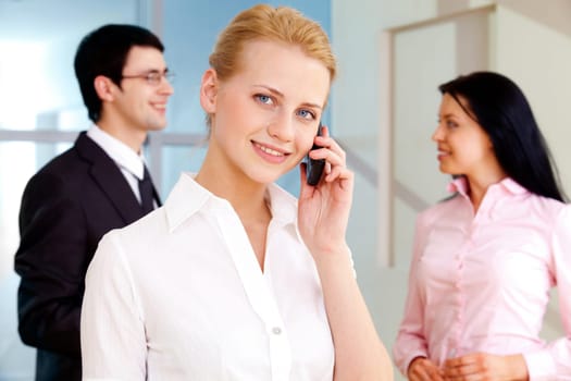 Image of pretty employee speaking on the phone and looking at camera in working environment