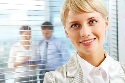 Female leader looking at camera with two partners interacting in office behind