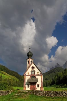 Small Church of St. John in Ranui with Odle Mountains and a dramatic cloudy sky,Funes Valley,Dolomites,Suedtitol,Italy