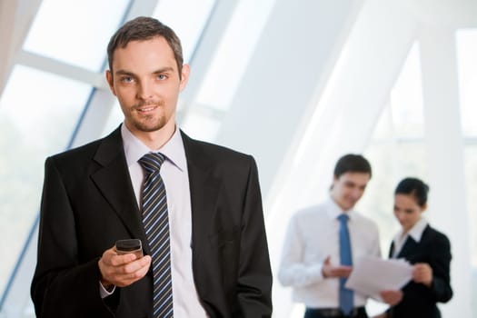 Portrait of successful businessman looking at camera with cell in hand on background of co-workers communicating with each other