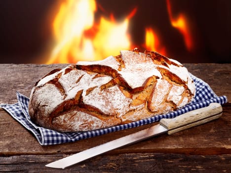 Crisp loaf of freshly baked rye bread with a knife on a checked napkin in front of a flaming fire