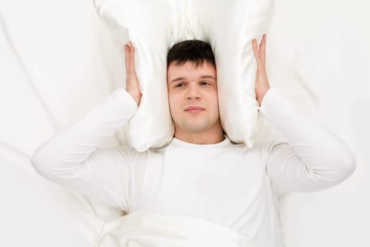 Tired man with his ears shut by a pillow and his eyes open lying in bed and trying to sleep