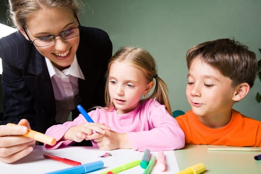 Image of smiling teacher holding highlighter and looking at girls picture while cute boy expressing his opinion
