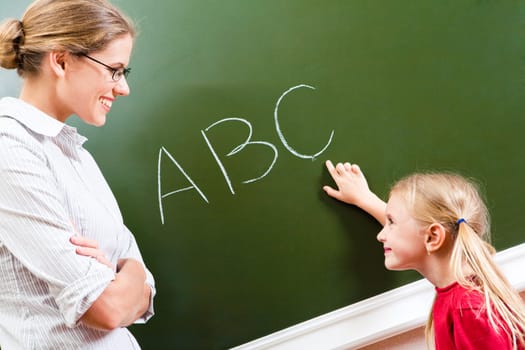 Image of smart girl pointing at letter on blackboard and looking at her teacher with smile