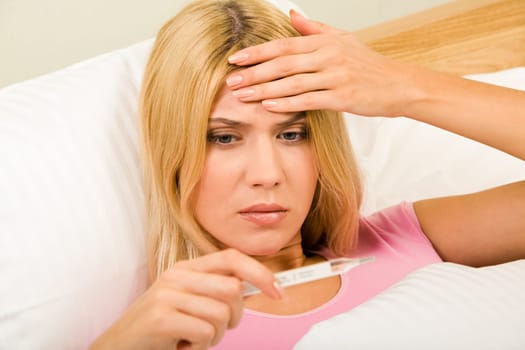 Photo of young woman lying in bed and looking at thermometer reading