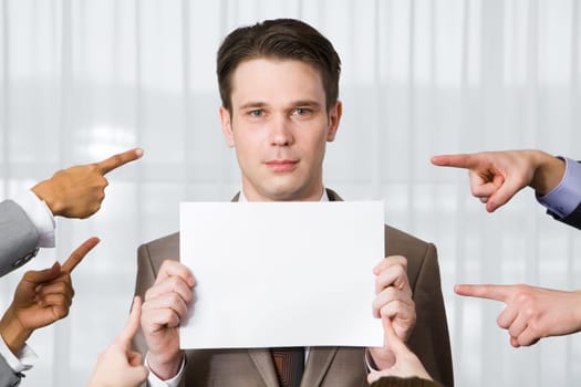 Serious businessman with blank paper looking at camera surrounded by pointing hands