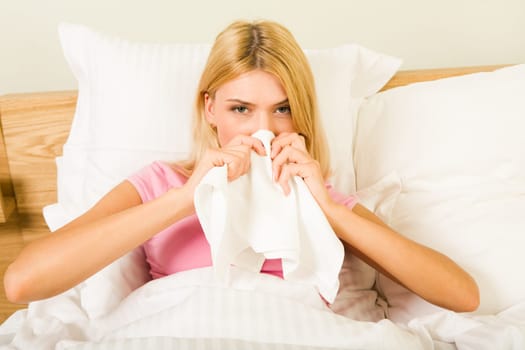 Photo of sick woman sitting in bed with tissue and blowing her nose into it