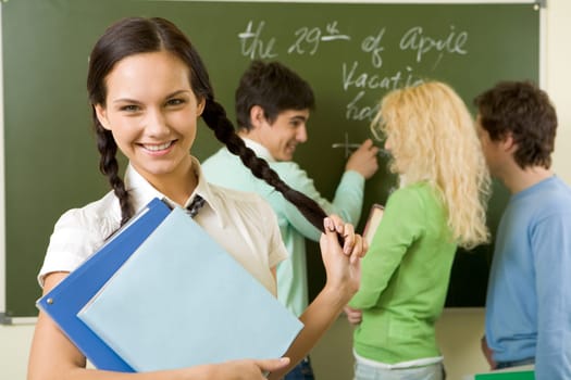 Portrait of cute girl holding textbook in hands on background of communicating students
