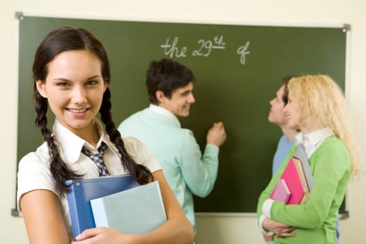Portrait of cute girl holding textbook in hands on background of communicating groupmates