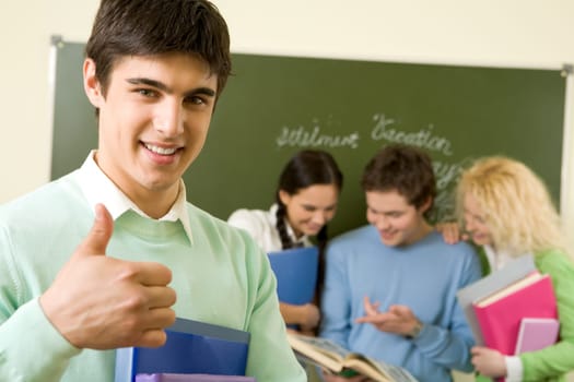 Portrait of cheerful lad showing sign of okay and looking at camera on background of reading groupmates