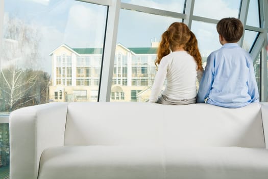 Rear view of two kids sitting on white sofa and looking through window