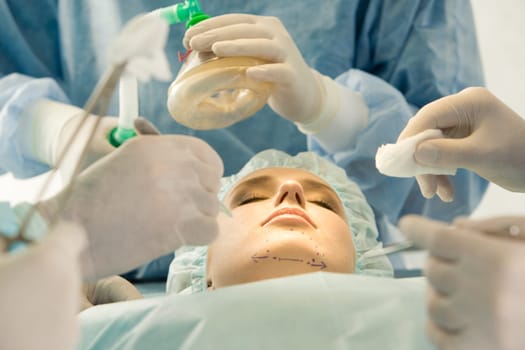 Photo of young female patient lying with closed eyes before operation with hands of doctors over her