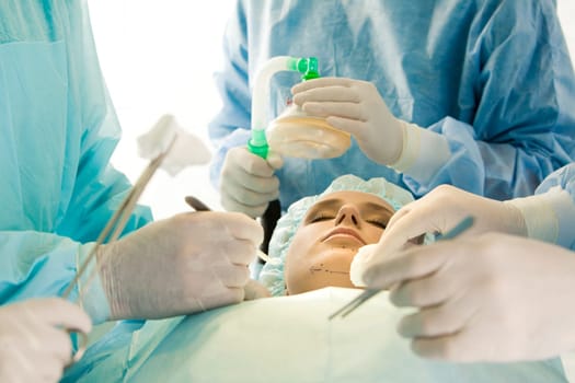 Photo of young female patient with closed eyes before operation with hands of surgeons over her holding surgical instruments and oxygen respirator