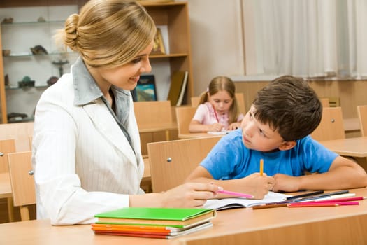 Photo of teacher and schoolboy sitting together at the desk and interacting during lesson