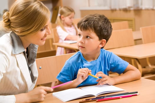Photo of teacher and schoolboy sitting together at the desk and interacting during lesson