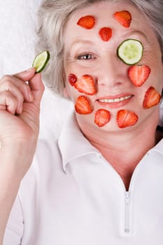 Aged woman holding cucumber slice by her eye and looking at camera