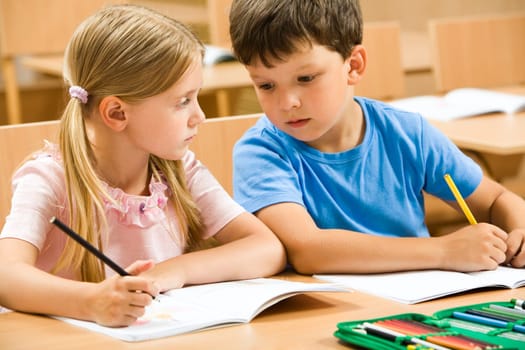 Portrait of two schoolchildren sitting by one desk during drawing lesson while boy looking at his mates copybook and girl watching at him