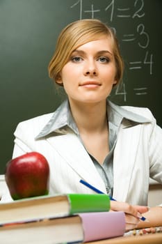 Portrait of confident teacher sitting at her desk with stack of books and apple on its top looking at camera