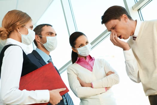 Group of co-workers in protective masks looking strictly at sneezing man