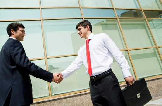 Image of two confident males handshaking outdoors at background of modern building