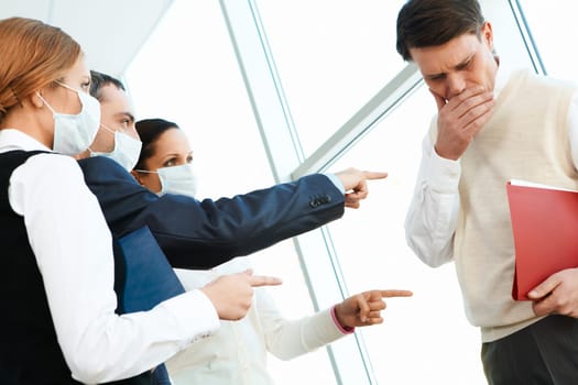 Group of associates in protective masks pointing at coughing man