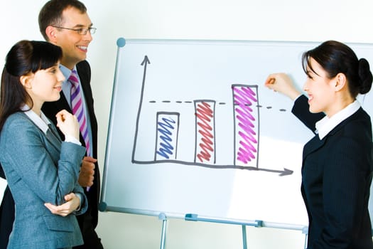 Photo of young businesswoman showing something on whiteboard with smile while her colleagues looking at it