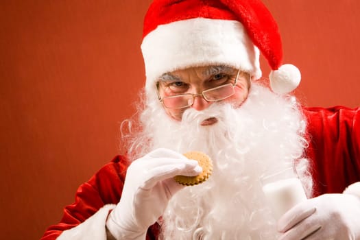 Photo of happy Santa Claus in eyeglasses looking at camera while drinking milk with bisquit