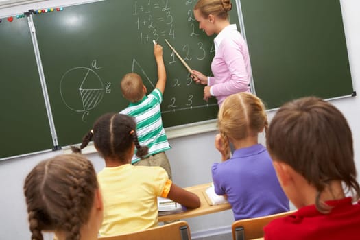 Photo of elementary student doing sums on blackboard and looking at it while his teacher helping him