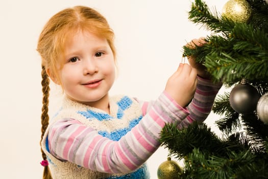 Photo of little girl near fir tree decorating it and looking at camera