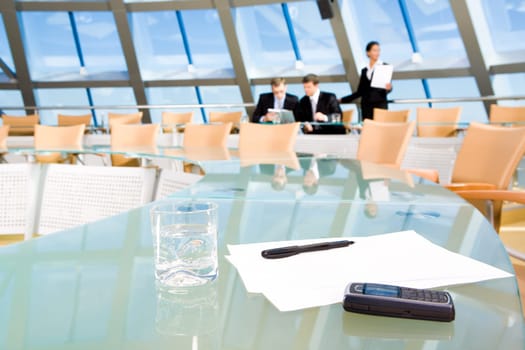 Image of blank papers, pen, cellular phone and glass of water on table of conference room