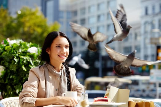 Image of happy female in open air cafe having coffee with cake in urban environment