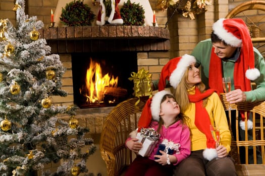Portrait of happy family in santa caps sitting by fireplace and looking at each other