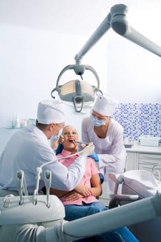 Image of young woman sitting on chair during oral sanitation surrounded by dentist and nurse