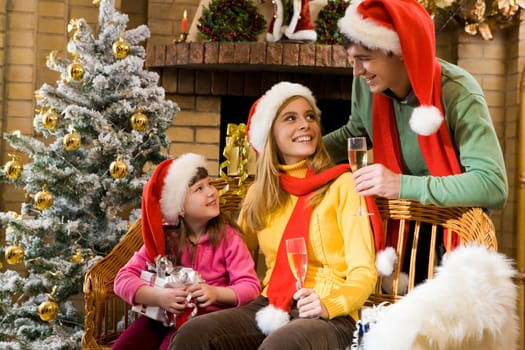 Portrait of happy family in santa caps sitting by fireplace and looking at each other on Christmas Eve